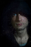 Portrait of a young hooded man behind glass covered in water drops - focus on the droplets