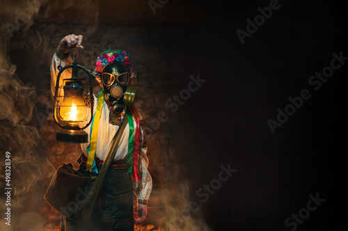 Ukrainian woman in a gas mask lights the way with an old gas lamp. Traditional folk costume. They hide in the basement, bomb shelters from chemical attacks, with poisonous gases. Copy space. © Sergey