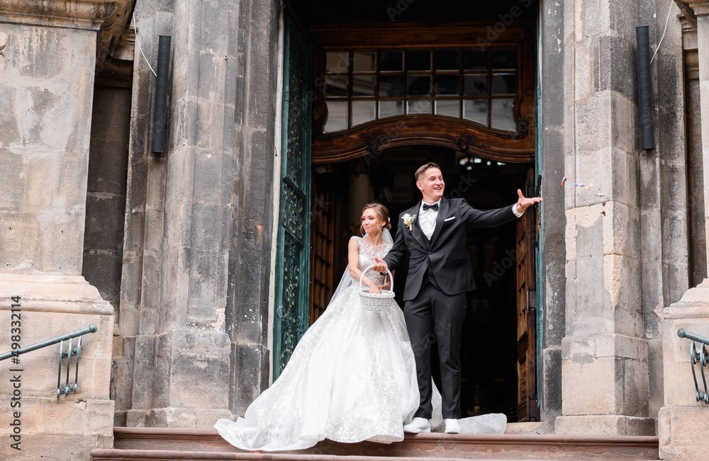 Front view of cheerful married couple, wearing in trendy wedding outfits, coming out from ancient building, stopped on centre of large wooden door, taking sweets from basket