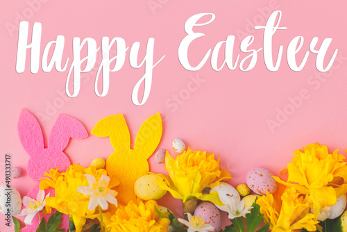 Happy Easter greeting card. Happy Easter text and colorful Easter bunnies, chocolate eggs and daffodils border on pink background, flat lay. Seasons greeting card, handwritten lettering