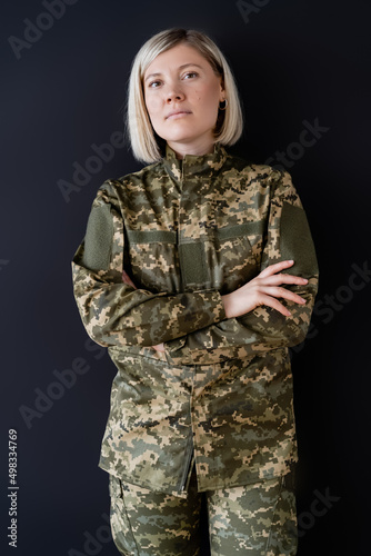 blonde military woman standing with crossed arms and looking at camera isolated on black.