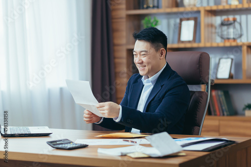 Young Asian man, businessman, satisfied director holding documents, letter, report, received good news. Sitting at a desk in the office, smiling