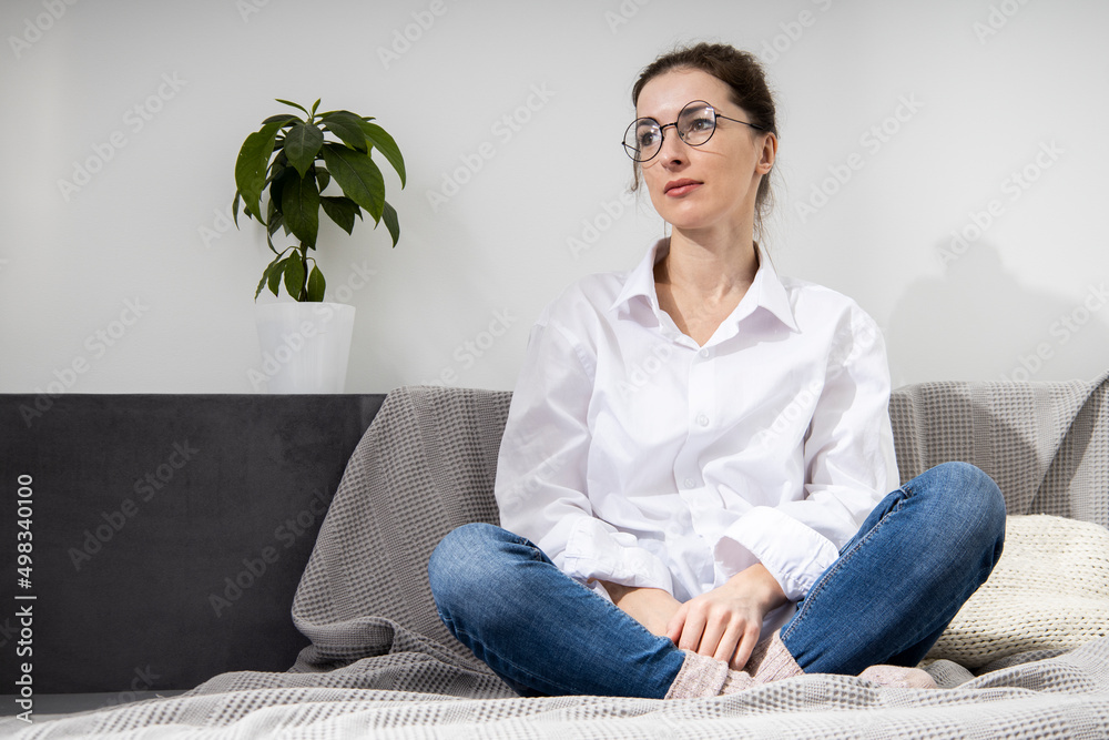 Thoughtful young woman in glasses in a white shirt sitting on a sofa. Thoughtful young woman in glasses in a white shirt sitting on a sofa.
