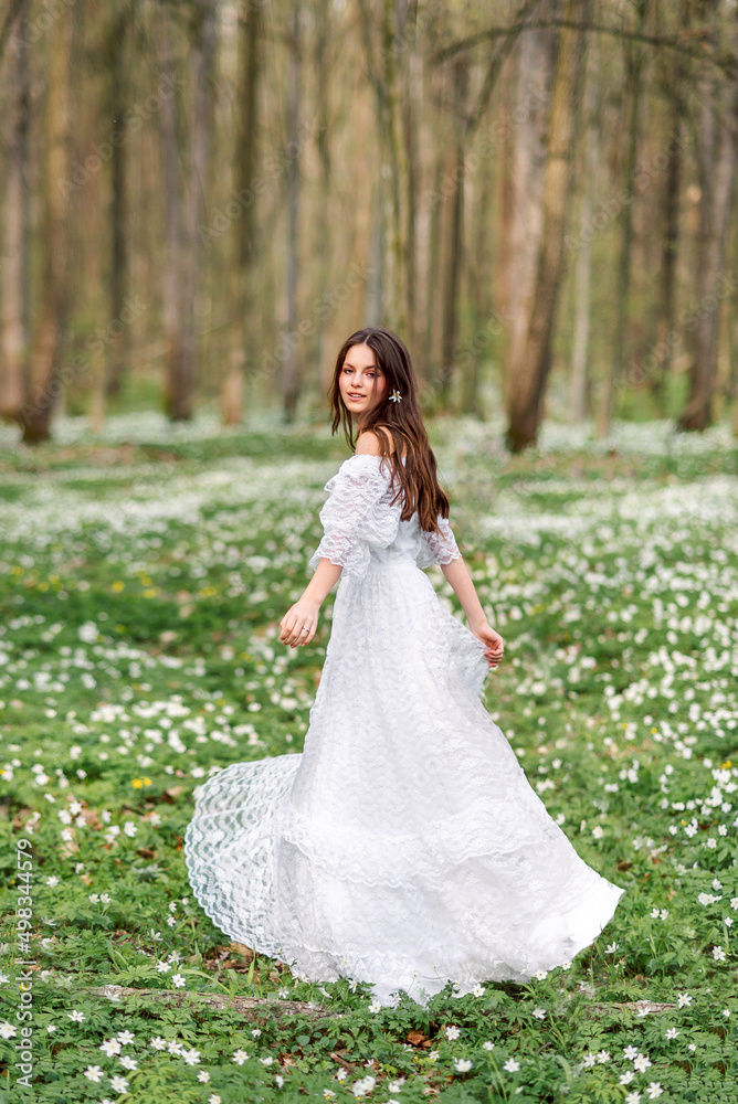 girl in a white long dress is spinning in a clearing with flowers. A young woman with long hair in a spring forest.