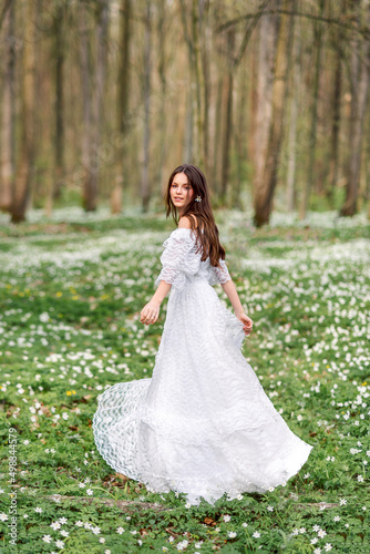 girl in a white long dress is spinning in a clearing with flowers. A young woman with long hair in a spring forest.