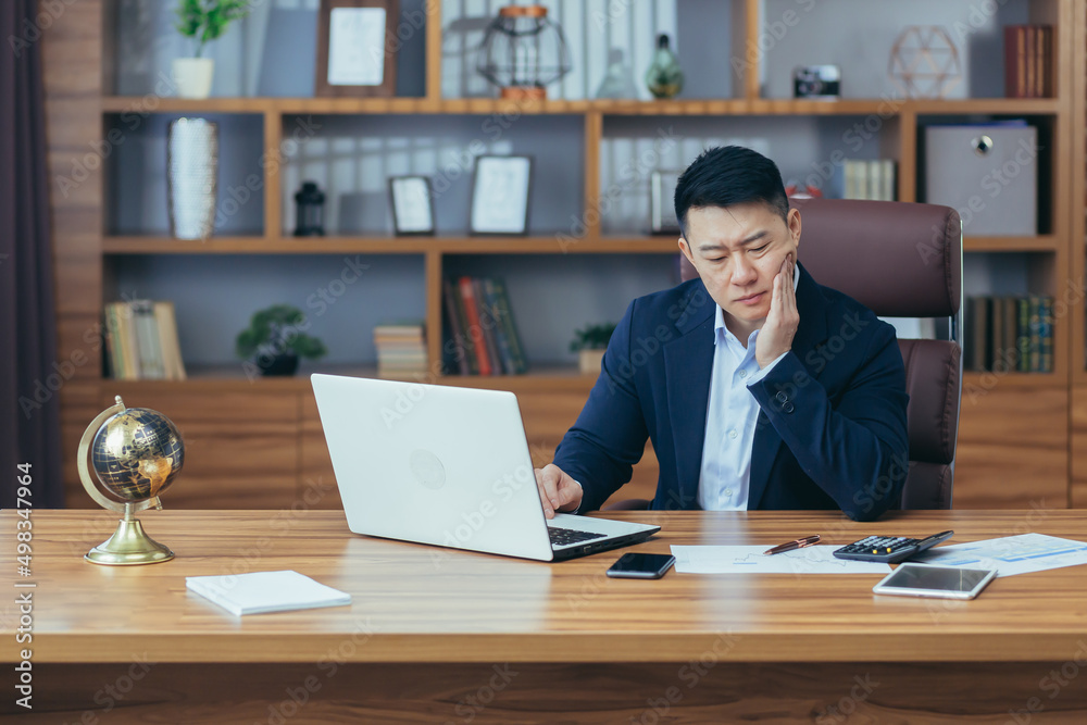 Toothache in the workplace. A young Asian man, a businessman, an employee, has a toothache at work. He is sitting at a desk in the office, holding his cheek, feeling pain Toothache in the workplace. A young Asian man, a businessman, an employee, has a toothache at work. He is sitting at a desk in the office, holding his cheek, feeling pain