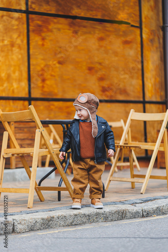 Stylish boy 3 years old in a leather jacket and brown trousers sits on the terrace near the cafe. Modern child. Children's fashion. Happy child