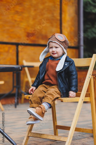 Stylish boy 3 years old in a leather jacket and brown trousers sits on the terrace near the cafe. Modern child. Children's fashion. Happy child © Дмитрий Ткачук
