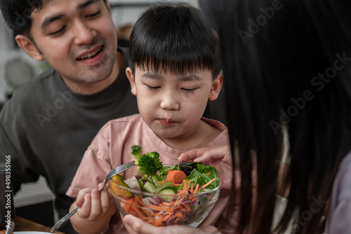Asian Cute little boy eating vegetable salad with his parents in the kitchen. Lovely Asian family having breakfast together. Happy family activity lifestyle concept.