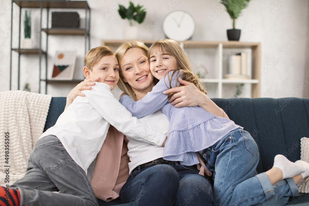 Happy little kids sit on couch hug cuddle excited young mom or nanny show love and care, overjoyed small children relax with mother embrace share sweet tender family moment in living room at home