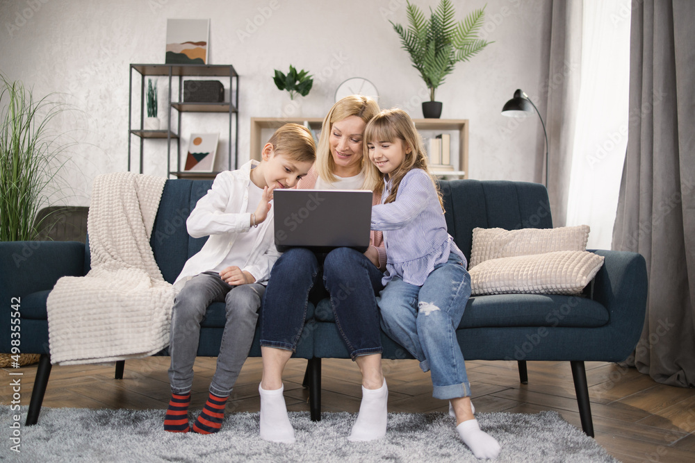 Happy family with two children having good time using laptop. Young smiling mother with cute daughter and son sitting on sofa at home looking at computer screen and watching video. Happy family with two children having good time using laptop. Young smiling mother with cute daughter and son sitting on sofa at home looking at computer screen and watching video.