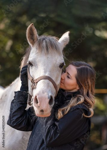 Young woman standing next to white Arabian horse eyes closed as if she's kissing or smelling, blurred trees background, closeup detail