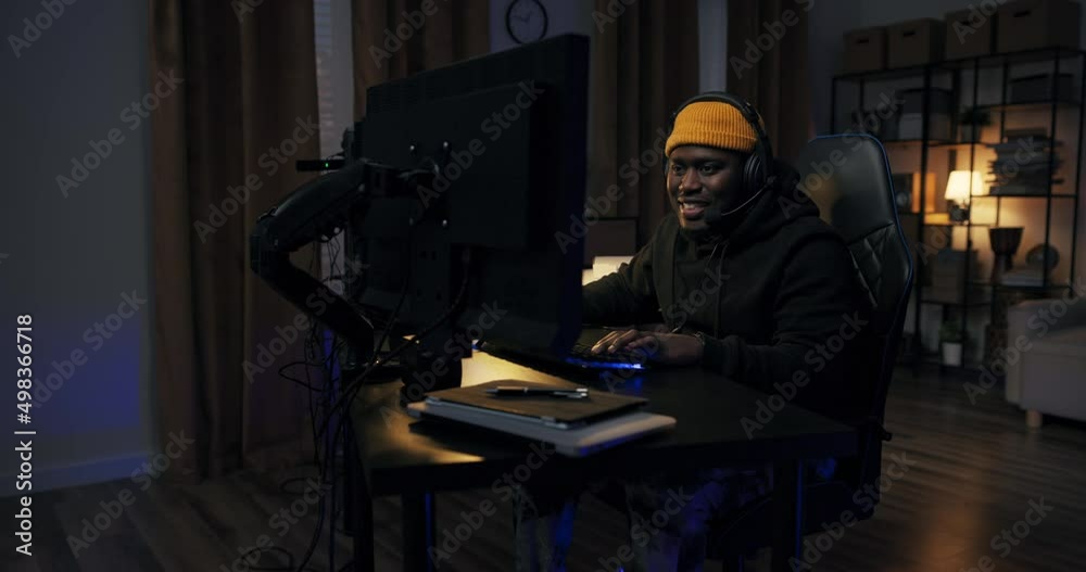 A man's success in a computer game. Boy in sweatshirt cap with headset raises hands up showing strength, victory, winning. He is smiling happy, hanging out in a dark room with led lights.
