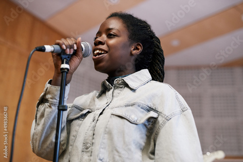 Low angle portrait of young black woman singing to microphone while recording music or rehearsing in professional studio