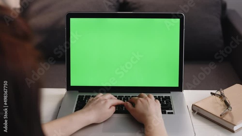 Rear view of a woman typing on the laptop with green screen, sitting at the wooden table. Female working at his desk at the computer, chromakey. Woman uses laptop with green mock-up screen.