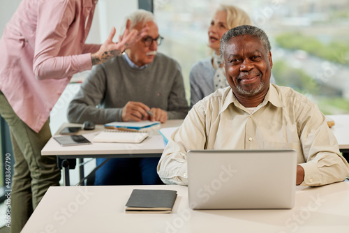 A senior african student smiling at the camera while group of multicultural senior students is discussing a programming project with an educator in the classroom. photo