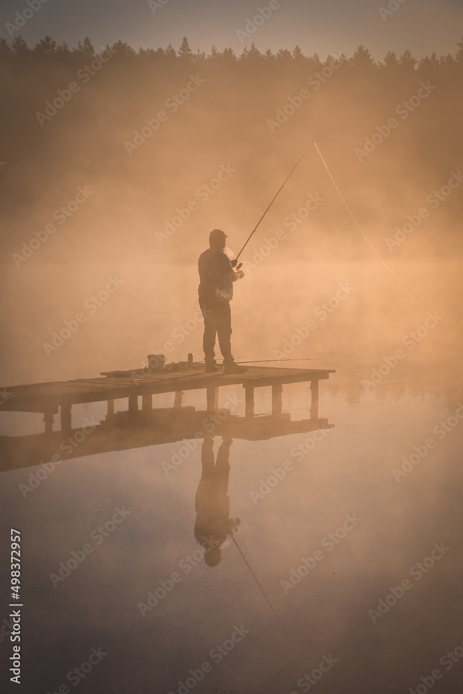 Beautiful morning scene by the lake. Silhouette of a man fishing on a wooden pier in the light of the rising sun.
