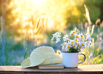 chamomile flowers in cup, book, braided rustic hat on table in garden, sunny natural abstract background. summer season. beautiful floral composition. relaxation, harmony atmosphere.