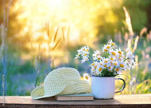 chamomile flowers in cup, book, braided rustic hat on table in garden, sunny natural abstract background. summer season. beautiful floral composition. relaxation, harmony atmosphere. photo