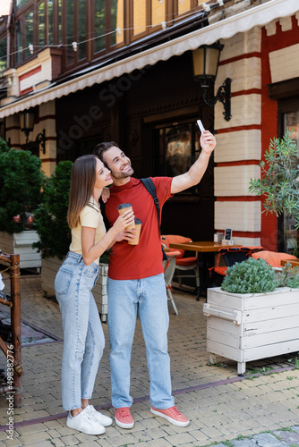 Positive couple with backpacks and coffee to go taking selfie on urban street.