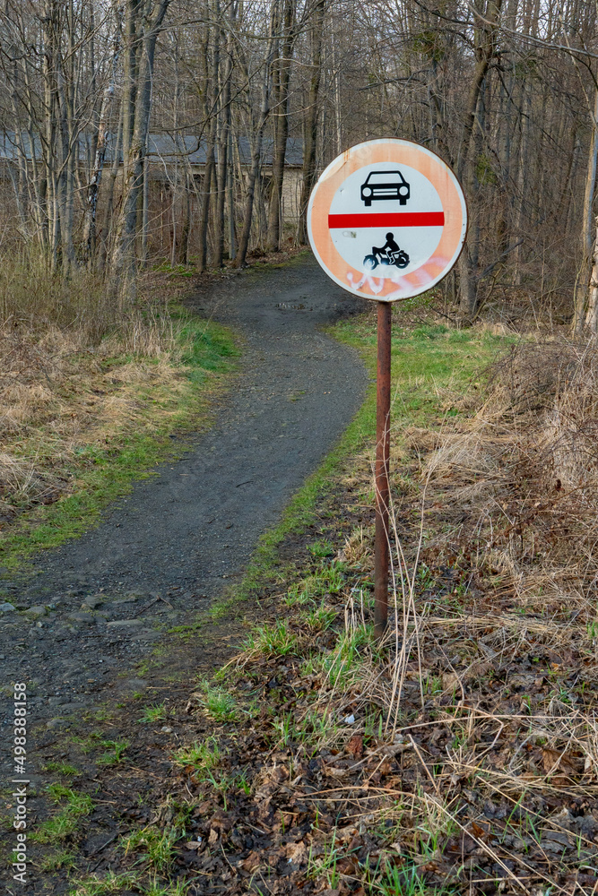 Naklejka premium No cars and no motorcycles sign next to a trail leading to a forest on a cloudy day of spring. Traffic sign in remote rural area.