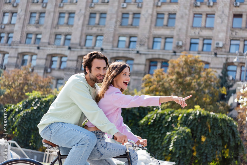 Excited woman pointing with finger while riding bike with boyfriend on urban street.