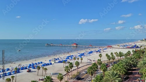 The pier of Clearwater Beach in Florida | A crowded Florida beach with people sunbathing and enjoying beach activities and seagulls flying | Also visible the palm trees and clouds in the horizon