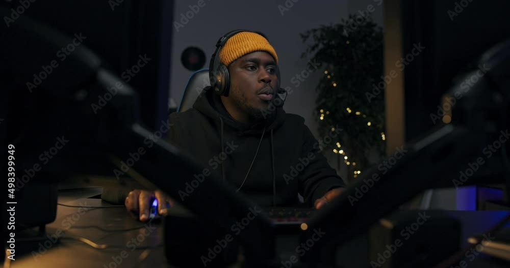 Men's room darkly lit with purple led lights, a portrait from behind a monitor of a man of American descent wearing a yellow cap with a headset. The guy is playing shooting games on a computer. Men's room darkly lit with purple led lights, a portrait from behind a monitor of a man of American descent wearing a yellow cap with a headset. The guy is playing shooting games on a computer.