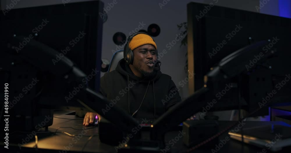 Dark room at night lit by led lights. Guy wearing cap headphones is sitting in comfortable chair in front of computer, playing games talking to friend, looking into camera with white teeth smile. Dark room at night lit by led lights. Guy wearing cap headphones is sitting in comfortable chair in front of computer, playing games talking to friend, looking into camera with white teeth smile.