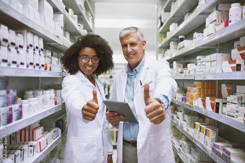 We are on track with stock. Portrait of two cheerful pharmacists holding a digital tablet and showing thumbs up while looking at the camera.