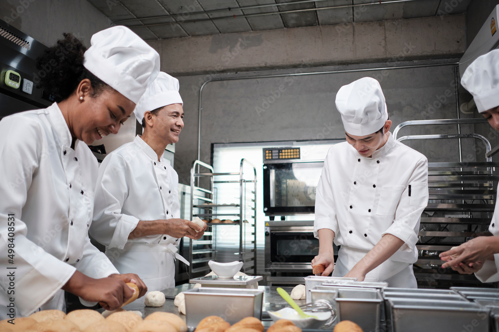 Multiracial professional gourmet team, four chefs in white cook uniforms and aprons knead pastry dough and flour, prepare bread, and bakery food, baking in oven at stainless steel restaurant kitchen.