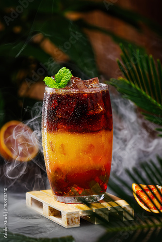 Bumble coffee with ice on a dark tropical background. Espresso, orange juice and syrup in layers in transparent glass with ice. Summer trendy refreshing coffee drink. Coffee house menu, recipe. Copy