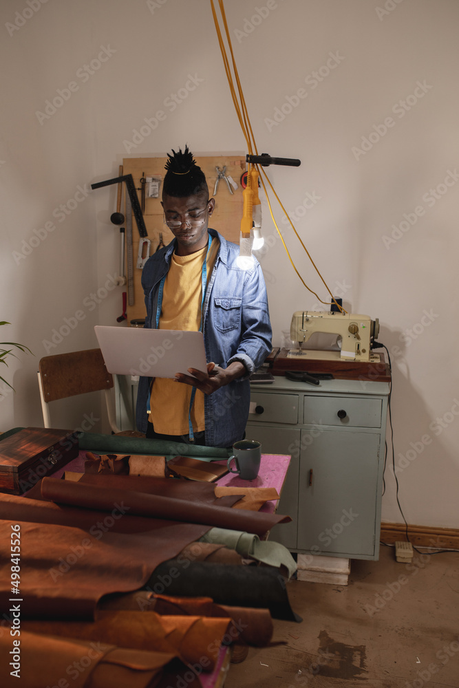 African american young craftsman using laptop while standing against wall in leather workshop African american young craftsman using laptop while standing against wall in leather workshop