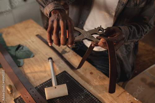 Midsection of african american young craftsman making hole on leather belt with punch plier photo