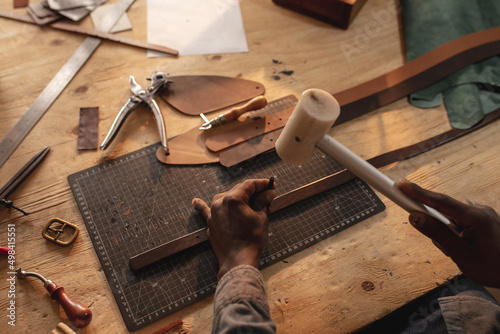 African american young craftsman making hole on leather belt with hammer and equipment photo