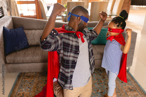 African american father and daughter wearing superhero costumes flexing muscles in living room photo