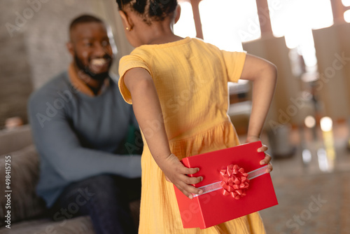 African american girl hiding surprise present behind her back for father in living room at home photo