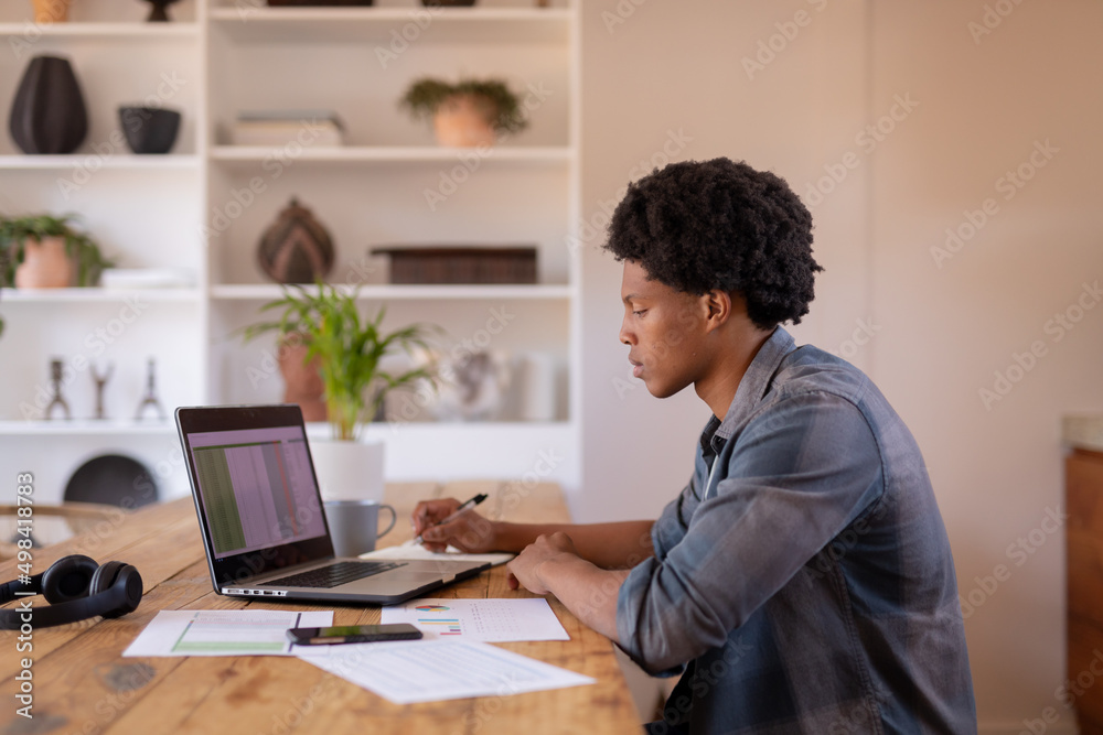 Side view of african american young businessman writing while using laptop at table Side view of african american young businessman writing while using laptop at table