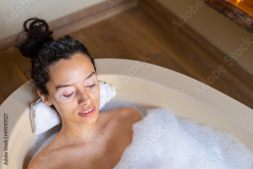 High angle view of biracial young woman with eyes closed relaxing in soapy bathtub at spa