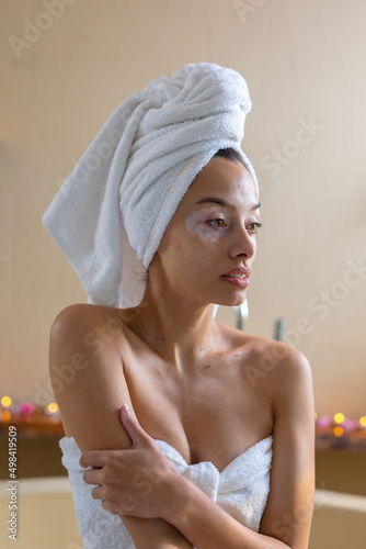 Thoughtful biracial young woman with towel wrapped on head and body sitting against wall in spa photo