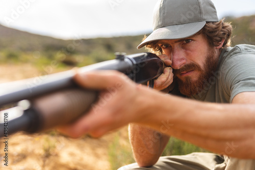 Confident young bearded caucasian man aiming rifle while hunting in forest photo