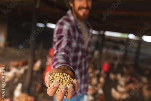 Smiling male poultry farmer showing grains in hand at pen with hens in organic farm photo