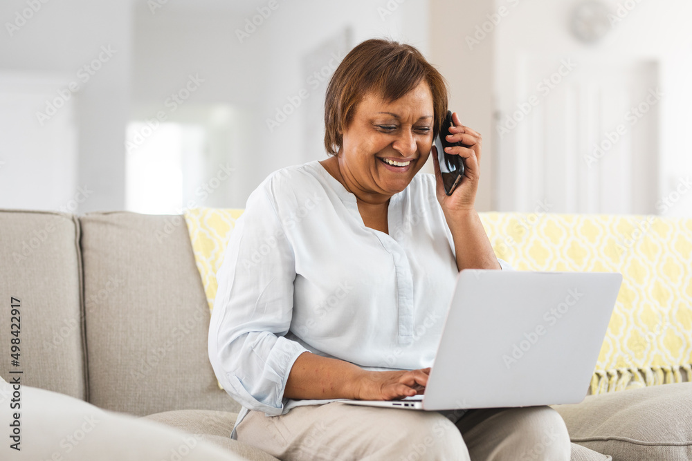 Happy african american elderly woman talking on smart phone while using laptop at home Happy african american elderly woman talking on smart phone while using laptop at home