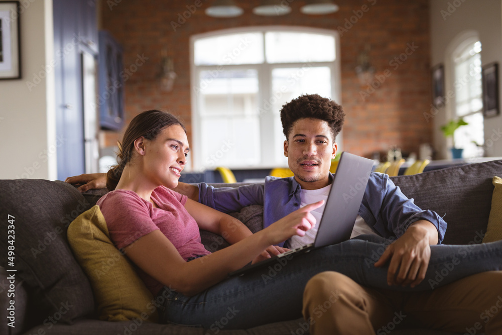 Young woman pointing at laptop while sitting with boyfriend on sofa in living room Young woman pointing at laptop while sitting with boyfriend on sofa in living room