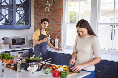Young man holding oil bottle talking to girlfriend chopping vegetables in kitchen at home