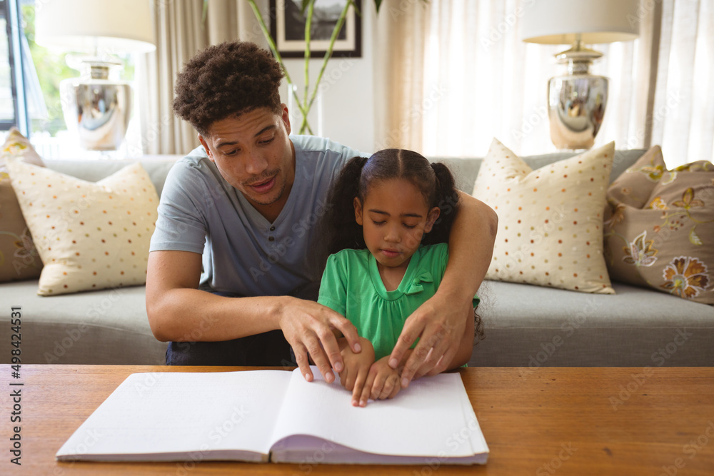 Biracial father helping blind daughter reading book while touching braille at home Biracial father helping blind daughter reading book while touching braille at home