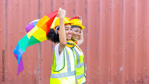 Young female African sisters in safety uniform and helmet show LGBT flag to support freedom and rights for gender equality next to red cargo or shipping container photo