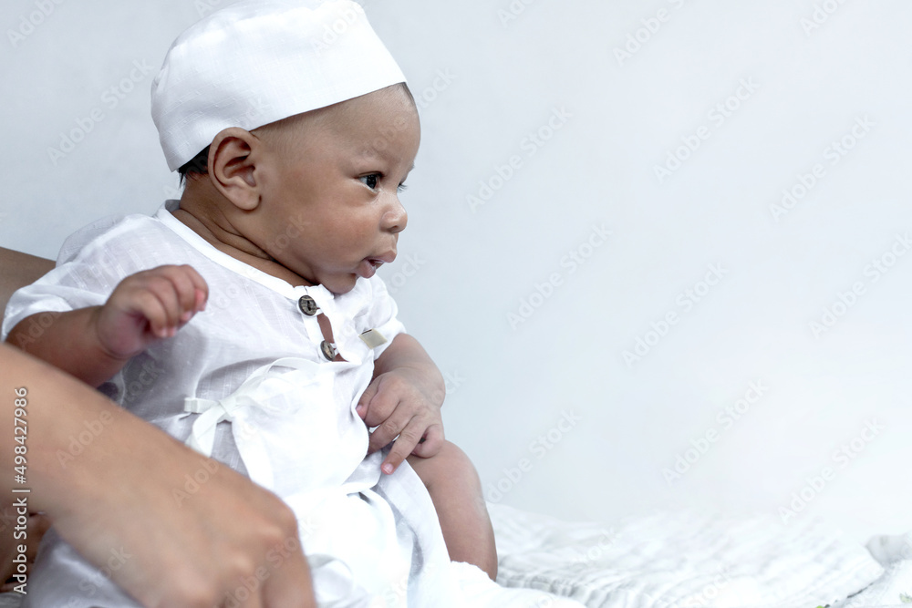 Cute 2 months old Muslim baby boy wearing white hat and Muslim Men's robe costume, isolated on white background, looking at something Cute 2 months old Muslim baby boy wearing white hat and Muslim Men's robe costume, isolated on white background, looking at something