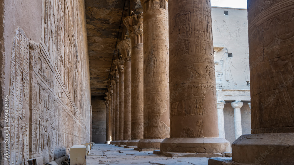 There is a narrow passage between the colonnade and the wall of the Temple of Horus in Edfu. Hieroglyphs, carvings on columns standing in a row are visible. Weathered stone floor and ceiling. Egypt