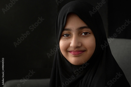 Close up portrait of little Muslim girl wearing black Hijab on black background, smiling at camera